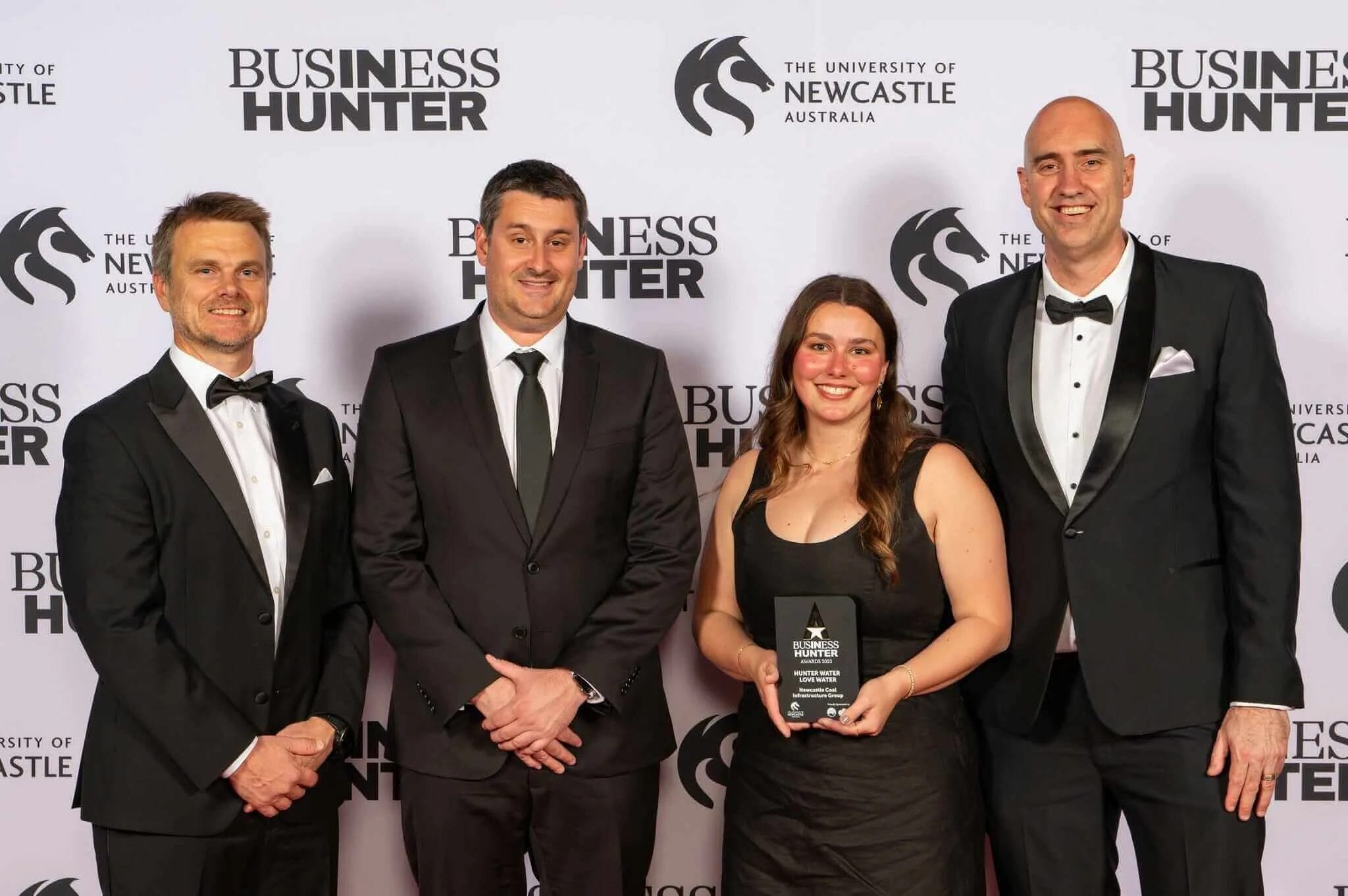 Three men in black suits and a lady in a black dress stand in front of a media wall with an award.